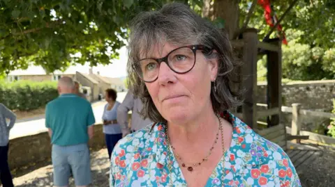A woman with dark hair and glasses, wearing a blue, white and orange floral top and a necklace, is pictured from the shoulders up in front of a tree, with more people gathered in the background behind her.