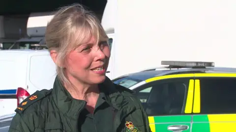 Jane Whichello - a blonde haired woman with a fringe and hair tied back wearing dark green ambulance attire, standing in front of an ambulance car.