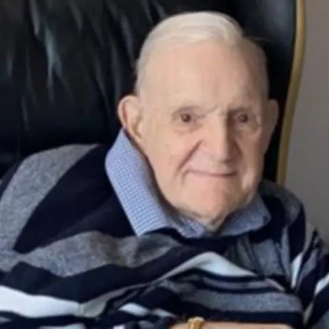 Family handout A man in his 80s with white hair grins at the camera. He is wearing a blue and white check shirt under a navy jumper.