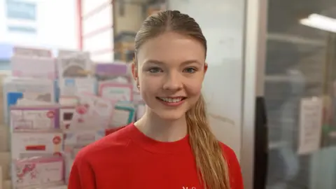 Elle is smiling at the camera. She is wearing a red sweatshirt and has long blonde hair tied in a ponytail. 