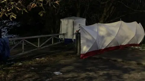 BBC A white police tent lies near shrubs on the edge of a path at the edge of Ashtons Field in the Little Hulton area of Salford. Close by, a white cover lies on top of a metal fence next to a gate.