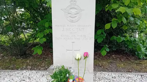 Nathalie te Wilt An image of a grave, the headstone reads 'Flight Sergeant J.MC C. Tait, Pilot, Royal Air Force, 26th March 1943, Age 21.' Two roses, one white and one pink and a plant have been placed at the bottom.