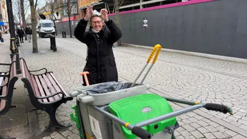 Ruth Major Ruth Major wearing a long black raincoat and glasses. She is standing on a public street beside wooden benches, holding up a piece of litter. In front of her there is a portable bin with litter picking tools.