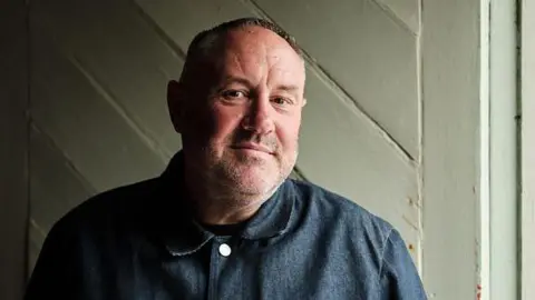 A man with short hair and a short beard standing in half-shade in front of a wooden door which is painted white and he is wearing a dark blue shirt.