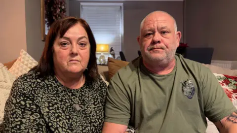 Elizabeth and Jonathan Osman looking at the camera. Elizabeth is on the right, she has dark hair and is wearing a black top with beige patter. Jonathan is on the left in a green t shirt.