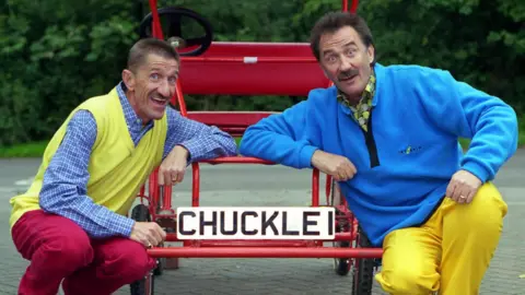 Two men kneel either side of a red two-seater pedal car with the number plate Chuckle 1