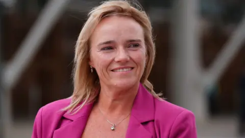 PA Media A smiling blonde woman wearing a pink blazer-style jacket outside the Scottish Parliament building, which is blurred in the background