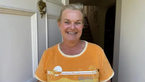 Photo of a woman with blonde hair tied back smiling at the camera in front of her front door. She is wearing an orange t-shirt.