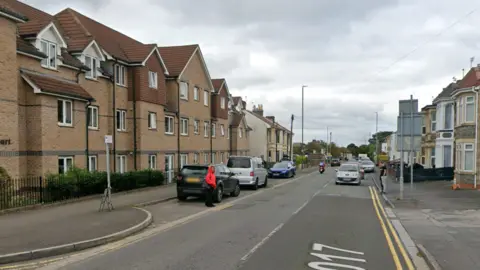Photo of North Street in Downend. There are three cars parked on the side of the road with a pedestrian wearing a red bright pink jumper. A silver car is driving on the road. 