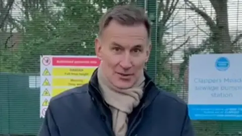 Godalming and Ash MP Jeremy Hunt pictured in front of the Thames Water Clappers Meadow sewage pumping station following the latest leak. He is wearing a navy coat and beige scarf.