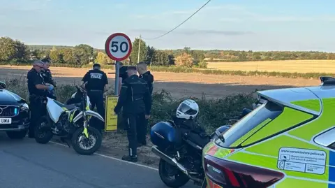 Warwickshire Police Police officers in black uniform seizing an illegal bike that they have seized. A 50mph sign can be seen near them and the rear of a police car to the far right of the picture.
