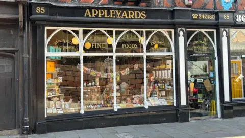 Charlie Stubbs / BBC There is a shop with big glass windows. The inner frames of those windows are white and the outer boarding is black. He has golden letters on the shop, on the top saying '85' , 'Appleyards' and '240180'. On the glass it says 'Appleyards Delicatessan, and 'Fine Foods.' 