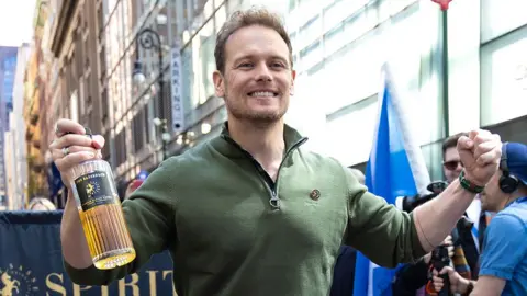 A man in a green top, smiling and holding a bottle of whisky in his hand, with a column of marchers behind him