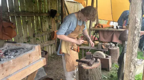 A man standing in an outdoor workshop. He is wearing a blue top, cream trousers and an apron. 