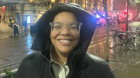 A woman wearing a black coat with the hood up is smiling. She wears a white shirt under the coat and has clear-framed glasses on. She is standing across the road from the Royal Exchange Theatre in Manchester city centre - visible in the background of the photo. 