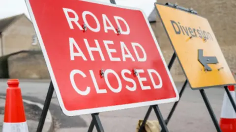 Kirklees Council A red road closure sign reading Road Ahead Closed stands next to a yellow diversion sign and traffic cones at a junction. Residential buildings can be seen in the background.