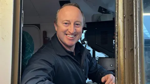 A train driver smiles into the camera. He is sitting in the driver's seat of a train. He is wearing a navy uniform.