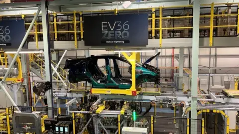 A production line at Nissan's factory in Sunderland. The shell of the New Nissan Leaf is central in the image. There are no car doors, wheels, front bumper or back bumper. It is a green colour.