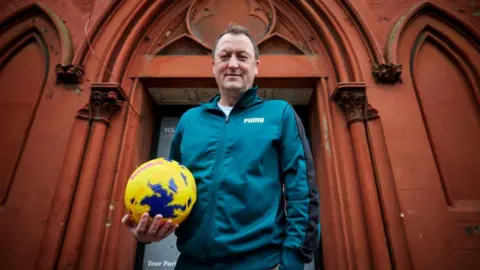 Jackson Lees Group Tony stands outside his community hub in Birkenhead with a football in hand. He is smiling and wears a teal green matching tracksuit.