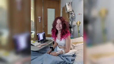 Millie Flemington-Clare Millie, aged about 18 with long curly pink hair. She is sitting upright on a hospital bed, smiling to camera. She has tubes coming out of her wrists