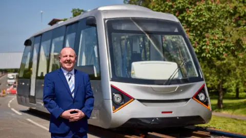 A bald man in a blue suit and stripy blue tie smiles at the camera while standing in the road in front of a very light rail vehicle, which is silver and looks like a tram on rails. It has a large front window with triangular lights on the front