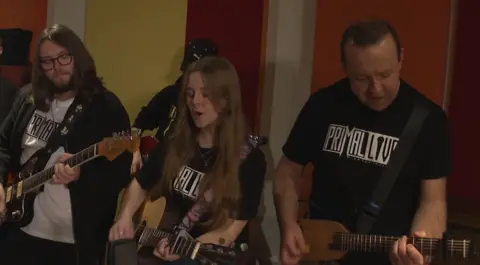 Three musicians playing guitar wearing black t-shirts with the Primal Live logo