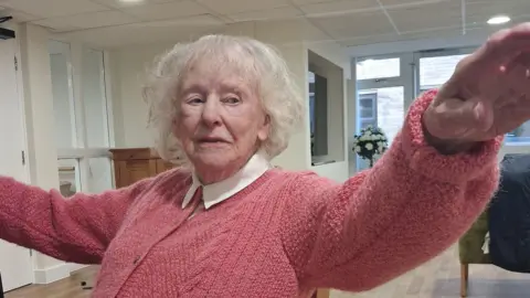 Josie demonstrates a dance move while sitting on a chair. Josie has short, white hair and a pink knitted cardigan with a white shirt colour visible. She extends her hands outwards.