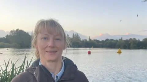 Clare Cowan/BBC A woman dressed in a warm brown jacket stands in front of the swimming lake