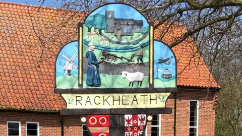 The village sign of Rackheath positioned outside. It is bright and colourful and features a pastor, a church, a sheep and someone ploughing.