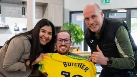 Jonny Butcher (centre) with his wife Ana (left) and head coach of Norwich City Philippe Clement (right). Jonny is holding a yellow and green football shirt showing the name BUTCHER above the number on the back.