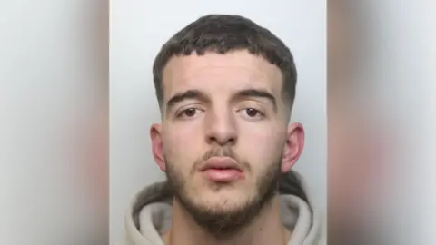 Rebian Bilalaj with short brown hair and beard staring at the camera and wearing a light-coloured hoodie in a police custody photo.