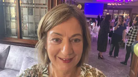 Alison Slater is standing in an events space with different tables set out and a blue projector screen. A sofa and chairs are visible to her left. She has fair hair, wears a silver chained necklace and has a white dress with brown flowers on it.