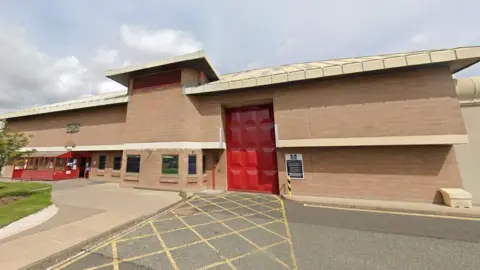 Front of Holme House prison. The low rise building is of brick, with a tall red door and a hatched area in front of it. To the left are some windows and what appears to be a security booth. 