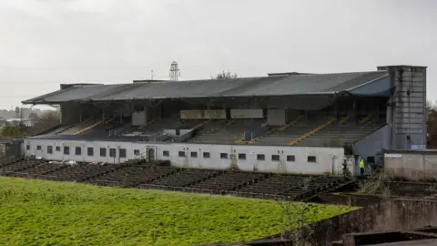 PA Media Dilapidated raised spectator stands at Casement park, overlooking a green pitch