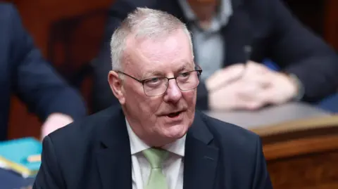A man with short grey hair is wearing glasses, a dark suit, white shirt and green tie. He is speaking at a conference