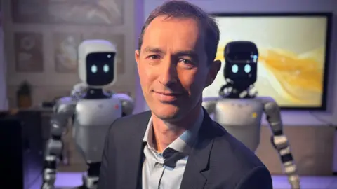Tim Ensor is wearing a blue suit and shirt. He is smiling at the camera and has a robot looking over each of his shoulders. The robots are humanoids - so have a head, body, arms and legs. Their heads are cubes with black screens for faces, displaying blinking white oval eyes. 