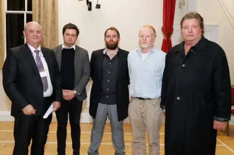 5 men stood posing for a photo in Alderney’s town hall. All in formal wear. From left to right,  Returning officer Theo Leisjer, Alex Snowdon, Iain Macfarlane, Stuart Clark, Edward Hill