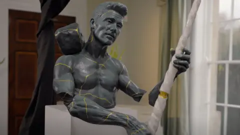 BBC/Chatterbox Media A grey sculpture replicating Darryn Frost's face, head and torso, crossed with gold lines. The sculpture is holding a broken white narwhal tusk and is mounted on a white plinth in a formal room.