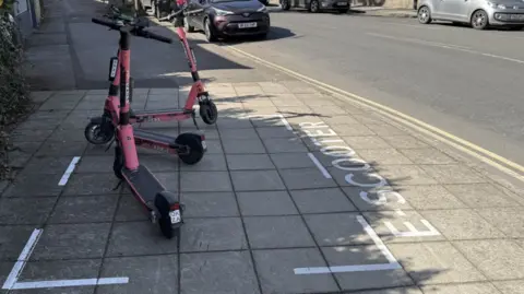Three red electric scooters parked inside a white parking bay space on the pavement which has the words 'E-SCOOTER' painted by it. 