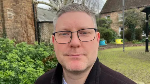 John Devine/BBC James Green has short grey hair and is in the middle of the image. He is wearing black framed glasses and a black coat. He is standing near houses.