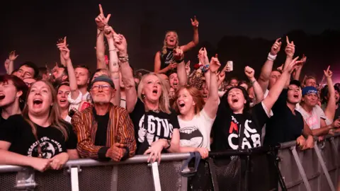 Front row of crowds at a concert, young and old scream and shout and look like they are having a great time
