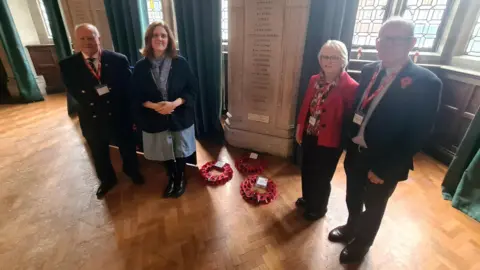 Dr Chris Oxley and his wife Ursula Oxley (right) with Principal Holly Bembridge and Archivist Kenneth Wright. They are standing at the memorial. There are poppies on the floor. A list of names can be seen as well as two more at the bottom. 