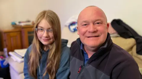 13-year-old Emily is sitting next to her father Alan in their sitting room