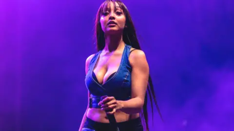 Getty Images PinkPantheress performs at Camp Flog Gnaw Carnival in LA in November 2023. She has long brown hair down her back and a fringe just past her eyebrows. She wears a cropped denim waist coat and holds a microphone in her left hand. She is lit by purple stage lighting