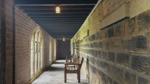 Hunters Estate Agents A corridor in a monastery with three benches. There is original brick stonework on the right-hand side and newer brickwork to the left. There are three lots of arched windows and black beams across the ceiling.