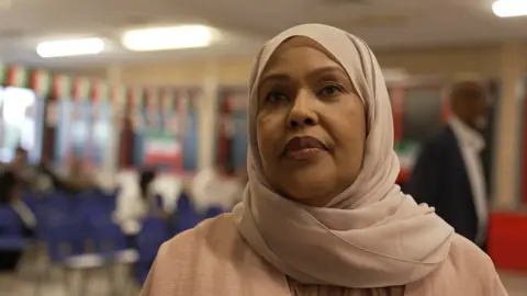 Fatima looks at the camera in a conference room. She's wearing a light pink head scarf and Abaya dress. 