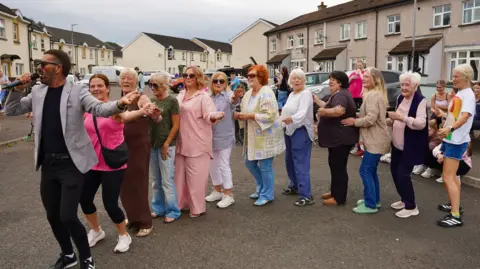 Derry Feile A sharply dressed man is holding a microphone while leading a line of women in a conga.