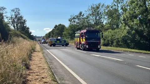 A road with trees on either side and several police and fire vehicles parked on it.