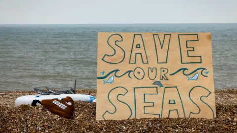 A handwritten sign on the beach at Hythe reads "Save our seas" as part of a protest organised by Surfers Against Sewage on 18 May 2024
