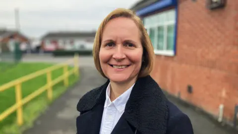 Woman with blonde cropped hear, wearing a navy coat with a large black collar and blue shirt, smiling in front of a bricked building and grass field with yellow railings.